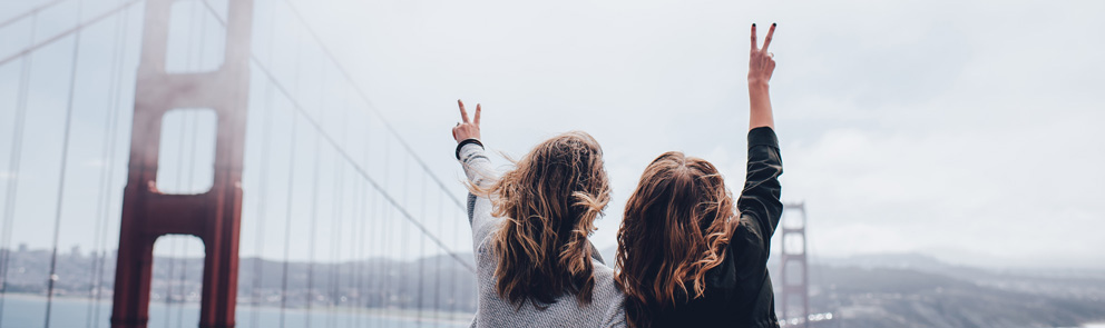 Bilde av to jenter som holder en arm opp hver og lager peace tegn. De ser på utsiket over Golden Gate bridge.